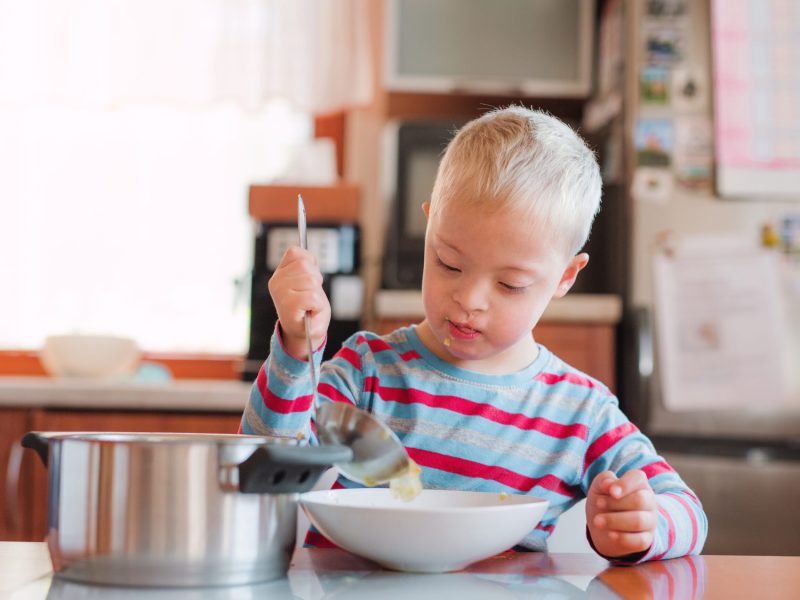 A handicapped down syndrome child pouring soup into a plate indoors before lunch.